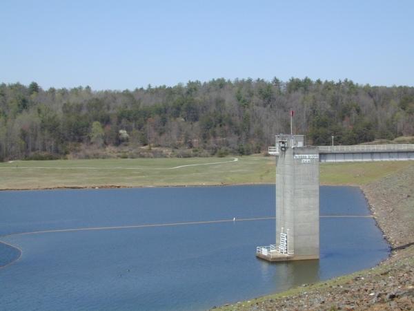 A concrete water tower stands near the edge of a calm lake, surrounded by a sloping shoreline and lush greenery. The background features a forested area with a mix of trees, indicating early spring or late winter. A clear blue sky is visible above, contributing to the serene scene. Dark Mountain Trail mountain bike trail.