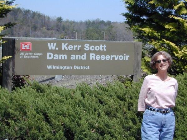 A woman standing beside a sign that reads "W. Kerr Scott Dam and Reservoir," which is part of the Wilmington District, US Army Corps of Engineers. The background shows trees and a clear sky, indicating an outdoor setting. Dark Mountain Trail mountain bike trail.