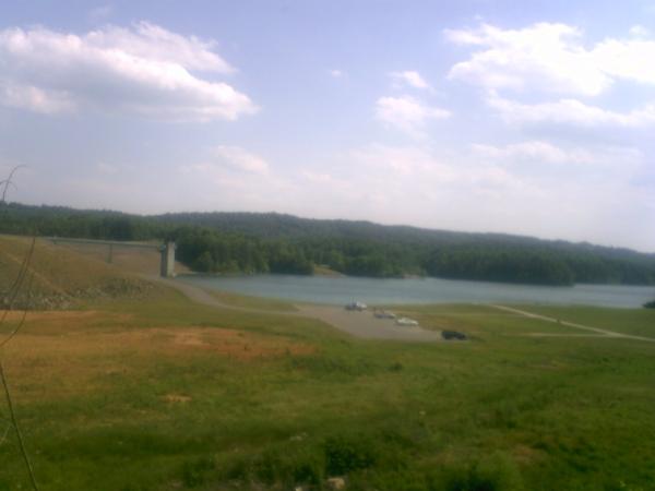A scenic view of a calm lake surrounded by green hills and a blue sky with scattered clouds. In the foreground, there is a patch of grassy land leading to the water's edge, with a few parked vehicles visible. In the distance, a bridge spans the lake, connecting two sides of the landscape. Dark Mountain Trail mountain bike trail.