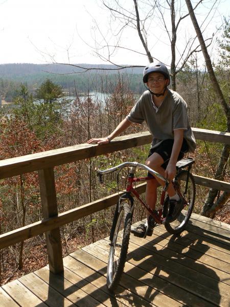 A person wearing a helmet sits on a red bicycle on a wooden platform, overlooking a scenic view of trees and a body of water in the background. The scene is bright and sunny, with sparse foliage indicating early spring or late fall. Dark Mountain Trail mountain bike trail.