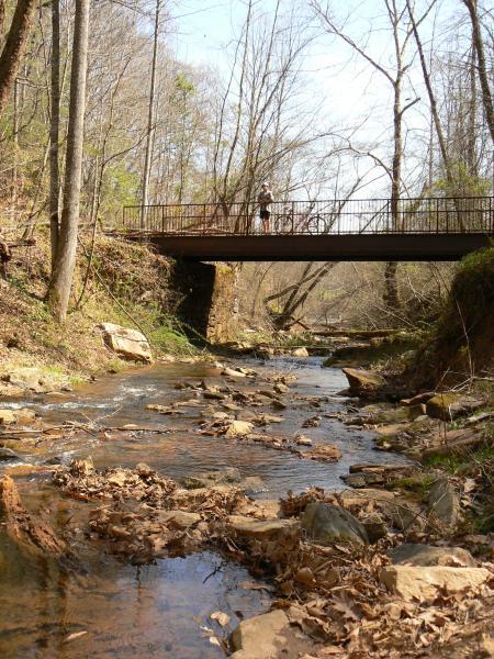 A serene natural landscape featuring a shallow stream with smooth stones and scattered leaves in the foreground. In the background, a metal footbridge crosses over the stream, surrounded by bare trees and lush greenery. The scene is bathed in natural sunlight, evoking a peaceful outdoor setting. Dark Mountain Trail mountain bike trail.