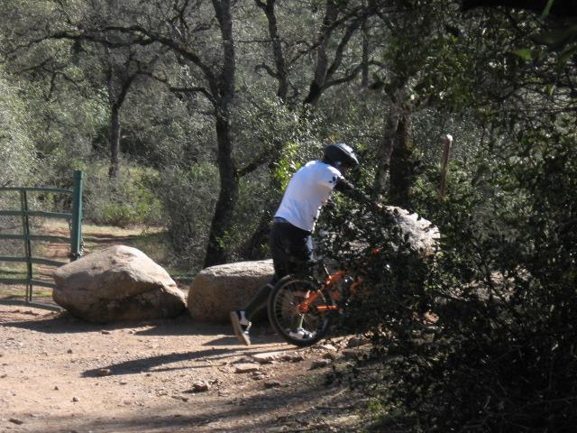 A person in a white shirt and black pants adjusts their mountain bike near large rocks on a dirt trail, surrounded by trees and vegetation. A metal gate is visible in the background. Clementine Loop mountain bike trail.