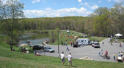 A scenic view of a large outdoor area featuring a pond surrounded by green trees. People are enjoying recreational activities, with tents set up and visitors walking and biking along the paths. Vehicles are parked near a gathering area, suggesting a community event or festival. The sky is blue with some clouds. Greenbrier State Park mountain bike trail.