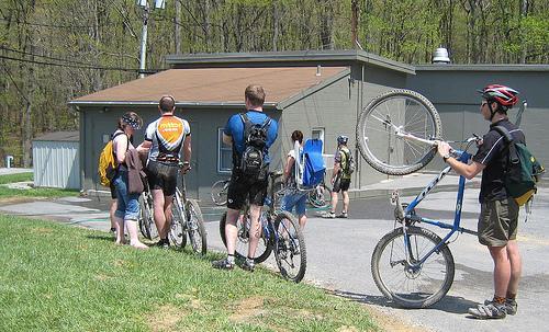 A group of five cyclists stands in line outside a building, preparing for a biking event or activity. They are wearing cycling gear, and some have backpacks. One cyclist is holding a mountain bike upside down, showing its wheels, while others wait near the entrance of the building. The setting features greenery and is likely in a park or recreational area. Greenbrier State Park mountain bike trail.