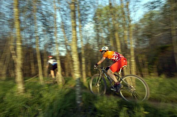 Two mountain bikers riding through a wooded trail at dusk. One cyclist, wearing an orange and red jersey, is in the foreground, captured in motion, while the second cyclist in the background is dressed in a white jersey. The scene is filled with trees and lush greenery, conveying a sense of speed and adventure. Greenbrier State Park mountain bike trail.