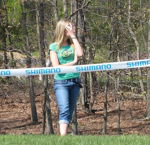 A young woman stands beside a Shimano-branded barrier in a wooded area, wearing a green shirt and denim jeans. She appears to be brushing her hair back while looking away from the camera, surrounded by trees and fallen leaves. Greenbrier State Park mountain bike trail.
