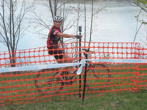 A cyclist wearing a helmet and a red and black jersey sits on a mountain bike next to an orange safety barrier, with a body of water and bare trees in the background. Greenbrier State Park mountain bike trail.