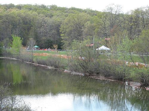 A serene landscape featuring a calm pond surrounded by lush green trees and shrubs. In the background, a small gathering area with tents can be seen, partially obscured by the trees, with a hint of orange fencing delineating the space. The still water reflects the greenery and the sky above, creating a peaceful outdoor scene. Greenbrier State Park mountain bike trail.