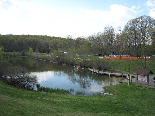 A serene landscape featuring a calm pond surrounded by lush greenery. In the background, trees are starting to bud, indicating early spring. A wooden walkway leads to a small dock at the water's edge, and there are tents set up in the distance, partially obscured by a fence. The sky is partly cloudy, creating a peaceful atmosphere. Greenbrier State Park mountain bike trail.