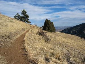 A winding dirt path leading through a grassy hillside with sparse trees, under a partly cloudy blue sky. The landscape features rolling hills and distant mountains. Mount Falcon Park mountain bike trail.
