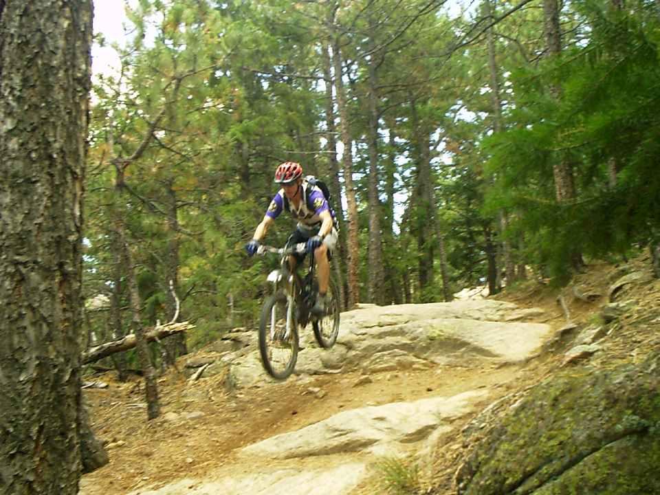 A mountain biker riding down a rocky trail in a forested area. The cyclist wears a helmet and biking gear, and is in motion, navigating over the rough terrain surrounded by trees. Mount Falcon Park mountain bike trail.