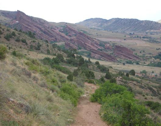 A scenic view of a winding dirt path leading through grassy terrain, surrounded by green shrubs and vegetation. In the background, striking red rock formations rise prominently against a backdrop of rolling hills and a cloudy sky. The landscape conveys a natural, rugged beauty typical of mountainous regions. Mount Falcon Park mountain bike trail.