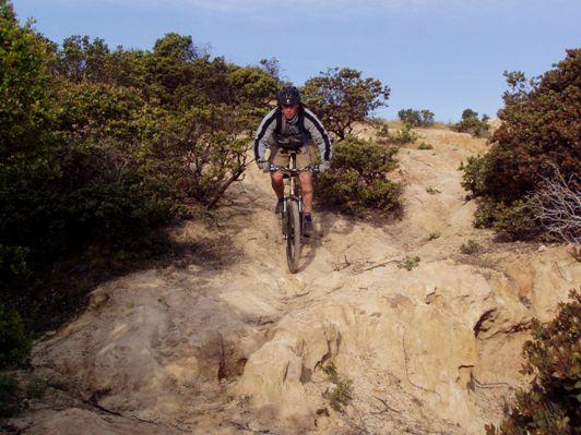 A mountain biker navigating a rocky trail, surrounded by greenery, with a clear blue sky in the background. The biker is focused as they descend a steep section of the trail. Fort Ord Public Lands mountain bike trail.