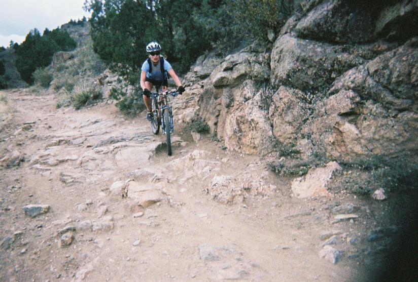 A person riding a mountain bike on a rocky trail surrounded by sparse vegetation and trees. The cyclist is wearing a helmet and outdoor gear, navigating through rough terrain with a mix of dirt and stones. Mount Falcon Park mountain bike trail.