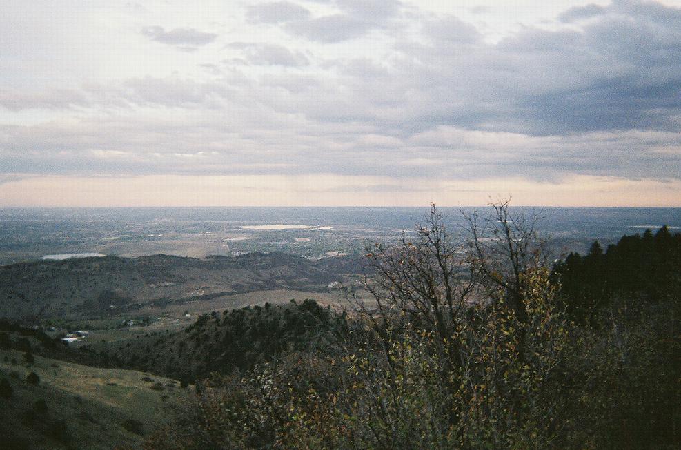 A panoramic view of a valley surrounded by rolling hills and mountains under a cloudy sky. The landscape features patches of greenery and hints of development in the distance, with a soft, muted color palette reflecting the overcast conditions. Mount Falcon Park mountain bike trail.