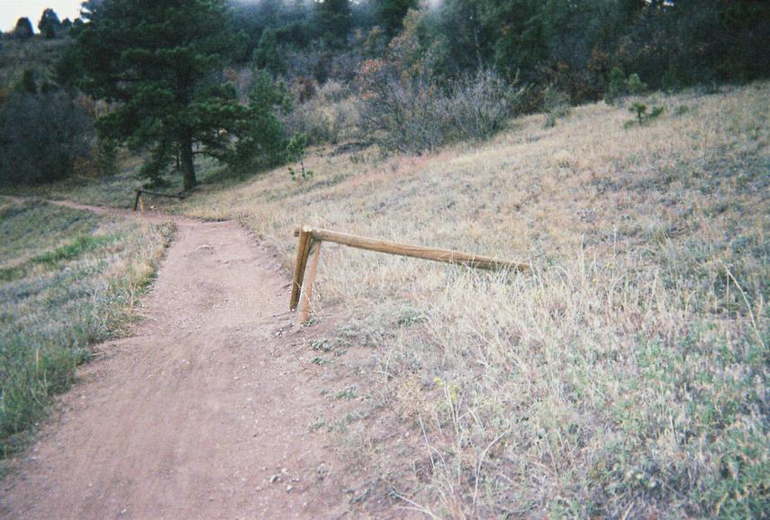 A dirt trail running through a grassy area, bordered by a wooden railing on one side. In the background, there are trees and shrubs, suggesting a natural outdoor setting. Mount Falcon Park mountain bike trail.