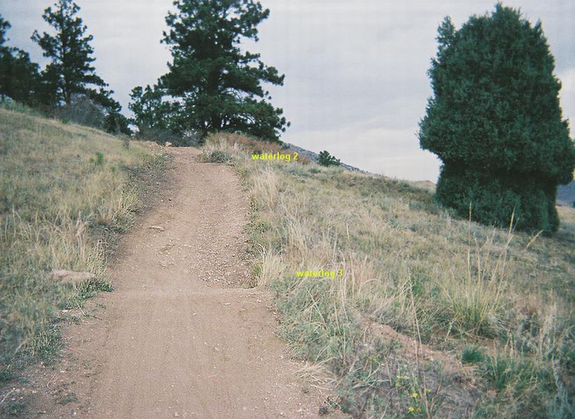 A dirt path leading through a grassy hillside, surrounded by scattered trees under a cloudy sky. The path appears slightly uphill, with markers labeled "waterlog 2" and "waterlog 3" visible along the side. Mount Falcon Park mountain bike trail.