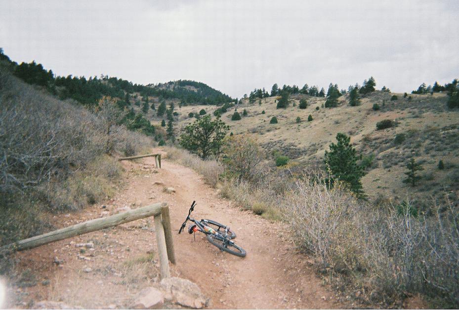 A mountain bike rests on a dirt trail surrounded by hills and sparse vegetation under a cloudy sky. The path curves between wooden posts, leading into the scenic landscape of rolling hills and distant trees. Mount Falcon Park mountain bike trail.