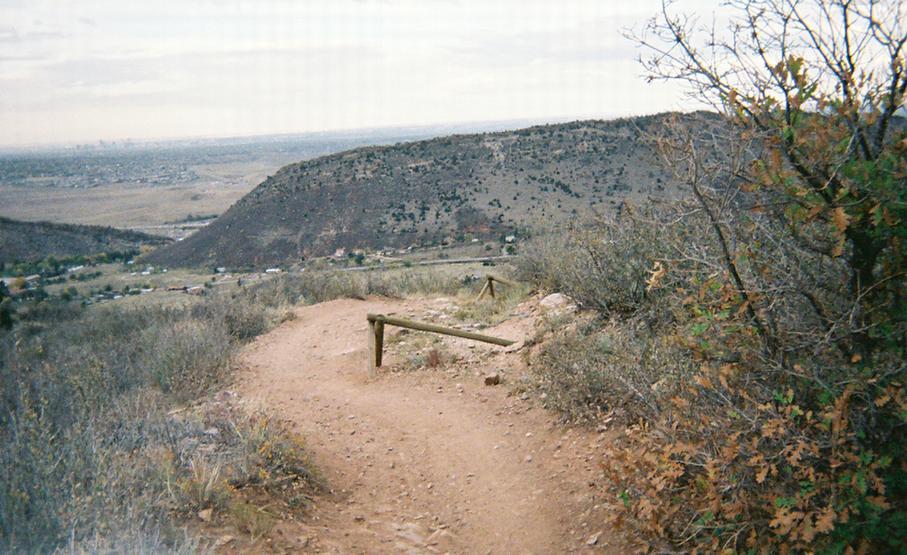 A winding dirt trail leads down a hillside, surrounded by sparse vegetation and shrubs. In the background, mountains rise against a cloudy sky, while a valley with small buildings and homes can be seen below. Mount Falcon Park mountain bike trail.