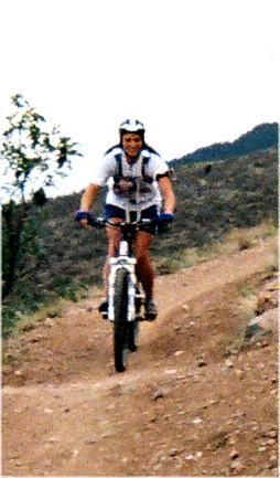 A person riding a mountain bike on a dirt trail, surrounded by hills and sparse vegetation. The cyclist is wearing a helmet and gear suitable for biking, and appears to be actively navigating the path. Mount Falcon Park mountain bike trail.