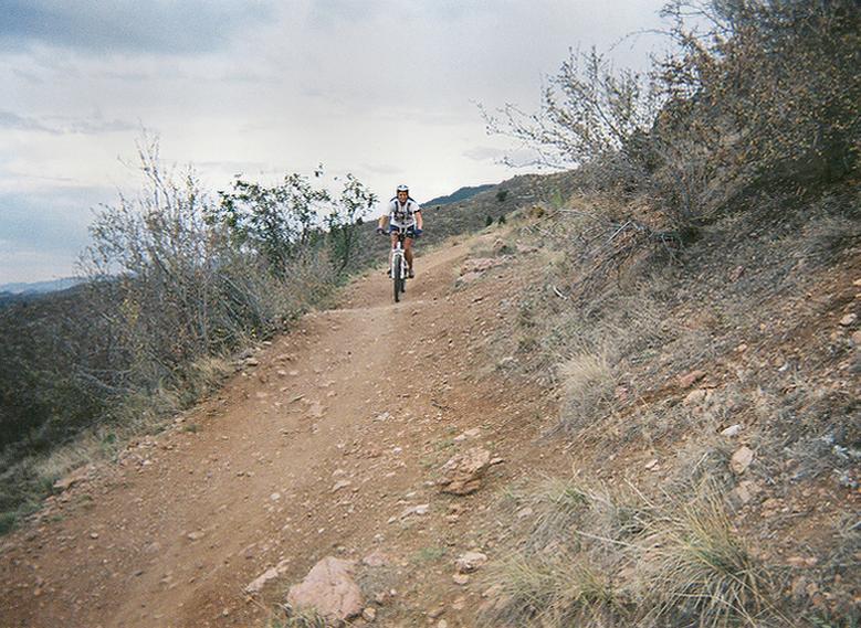 A person riding a mountain bike on a dirt trail surrounded by sparse vegetation and rocky terrain, with a cloudy sky in the background. Mount Falcon Park mountain bike trail.