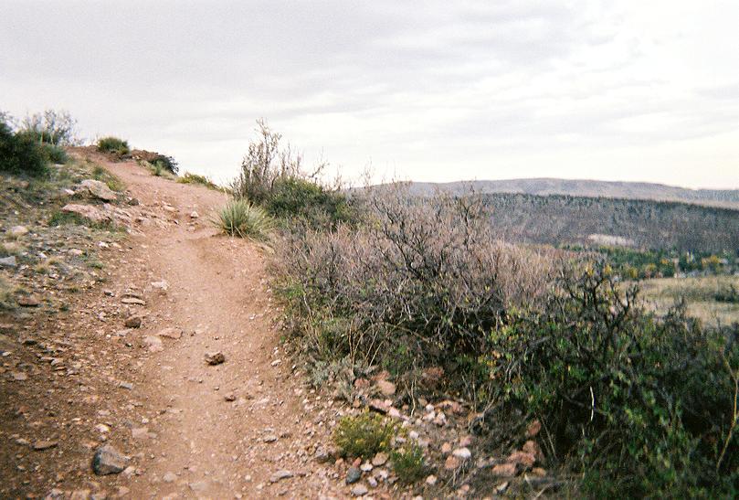 A dirt hiking path winding along a hillside, surrounded by sparse vegetation and rocky terrain. The sky is overcast, and the distant landscape features rolling hills and a valley, suggesting a natural outdoor setting. Mount Falcon Park mountain bike trail.
