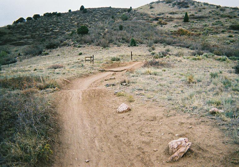 A dirt trail winding through a grassy hillside, with patches of shrubs and small rocks scattered along the path. In the background, there are rolling hills and a few trees. A fence can be seen along the side of the trail, indicating the boundary of the area. The sky is overcast, giving a muted tone to the landscape. Mount Falcon Park mountain bike trail.