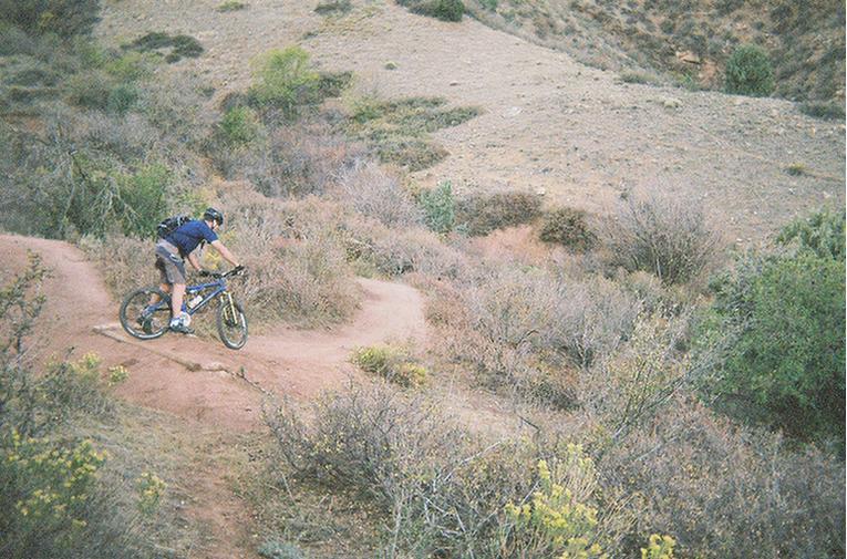 A mountain biker navigates a winding dirt trail surrounded by sparse vegetation and rolling hills. The scene captures the outdoor adventure spirit in a natural landscape. Mount Falcon Park mountain bike trail.