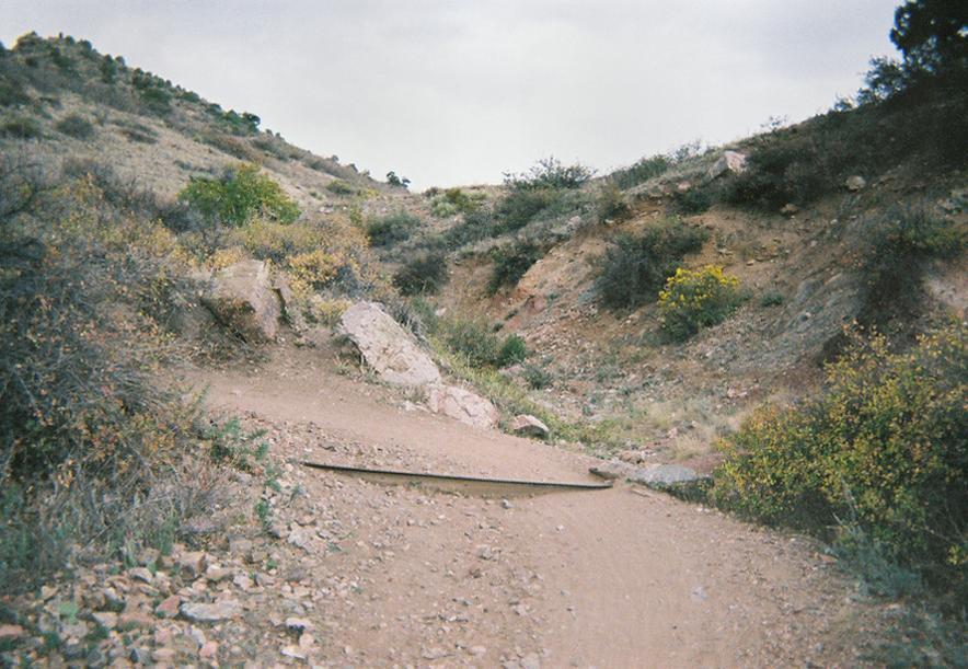 A dirt path winding through a hilly landscape covered in sparse vegetation, with rocks scattered along the sides. The sky is overcast with gray clouds, creating a muted light over the scene. Mount Falcon Park mountain bike trail.