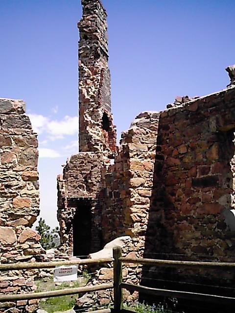 Ruins of a stone structure with a tall chimney, surrounded by partially collapsed walls. A wooden fence encloses the area, and a sign is visible in the foreground indicating "Restricted Access." The sky is clear and blue, with a few scattered clouds. Mount Falcon Park mountain bike trail.