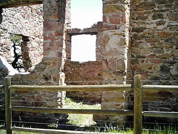 Alt text: View of the interior of a dilapidated stone building, with crumbling walls and a window opening. A wooden fence is visible in the foreground, and greenery can be seen through the building's openings. Mount Falcon Park mountain bike trail.