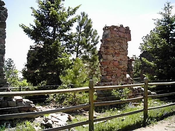 Ruins of an old stone structure surrounded by trees and greenery, with a wooden fence in the foreground. The sky is clear and sunny, highlighting the natural landscape. Mount Falcon Park mountain bike trail.