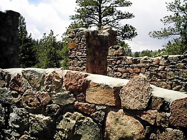 Alt text: Ruins of a stone structure with moss-covered walls, surrounded by tall pine trees and a blue sky with scattered clouds. Mount Falcon Park mountain bike trail.
