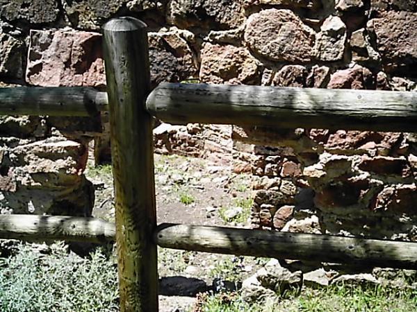 A wooden fence made of rounded logs in front of a weathered stone wall. The scene includes grassy ground and hints of vegetation around the fence, suggesting a natural outdoor setting. Mount Falcon Park mountain bike trail.