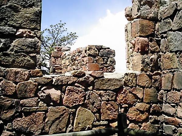 Ruins of a stone structure with exposed walls, featuring a scattering of stones in various colors. The scene is illuminated by natural light, with a glimpse of trees visible through the openings and a bright sky above. Mount Falcon Park mountain bike trail.