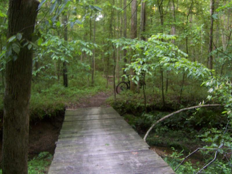 A wooden footbridge crossing a small stream, surrounded by lush green vegetation in a forest. In the background, a biking path is visible, with a bicycle partially obscured by trees. Canal Loop mountain bike trail.