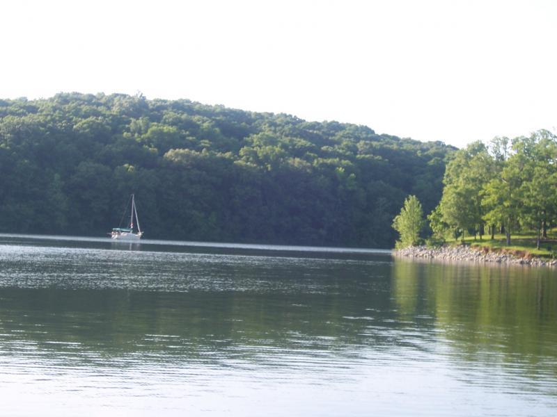 A tranquil scene of a serene lake surrounded by lush green hills. A small sailboat is gently anchored on the water, reflecting the landscape, while trees line the shore, creating a peaceful natural setting. Canal Loop mountain bike trail.