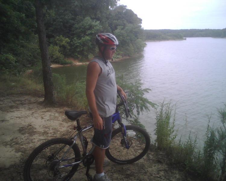 A person wearing a helmet and sunglasses stands next to a mountain bike beside a calm lake. The scene is surrounded by trees, and the shoreline is visible in the background. The atmosphere appears tranquil and outdoor-focused. Canal Loop mountain bike trail.