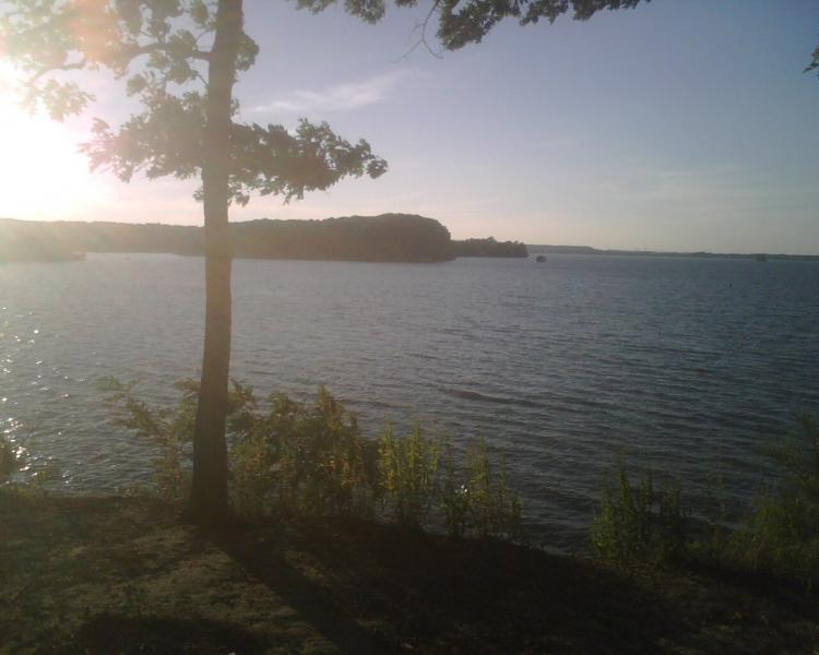 A serene lake scene at sunset, featuring gentle waves and a silhouette of trees along the shoreline. The sky is painted with soft hues, and the sun is low, casting a warm glow over the water. Canal Loop mountain bike trail.