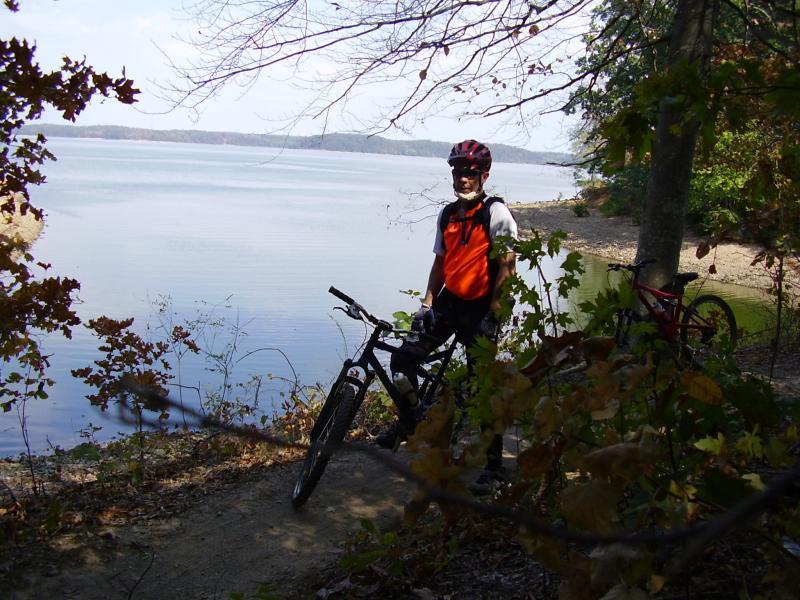 A cyclist in an orange shirt and helmet stands beside a mountain bike near a calm lake, surrounded by trees and autumn foliage. The scene captures a peaceful outdoor adventure on a sunny day. Canal Loop mountain bike trail.