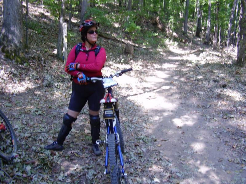 A person wearing a red long-sleeve shirt, black shorts, gloves, knee pads, and sunglasses stands next to a mountain bike on a dirt path in a forested area with trees and fallen leaves. Canal Loop mountain bike trail.