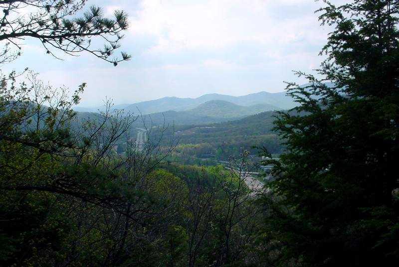 A panoramic view of rolling green hills and distant mountains under a cloudy sky, framed by trees in the foreground. The landscape showcases the beauty of nature, with varying shades of green and gray, suggesting a serene outdoor environment. Kitsuma mountain bike trail.