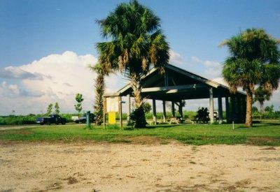 A shaded picnic pavilion surrounded by palm trees, with a grassy area and a dirt path in the foreground. In the background, a blue truck is parked, and there are white clouds in a blue sky. Caloosahatchee Regional Park mountain bike trail.