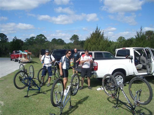 Group of cyclists in athletic gear adjusting their bikes in a grassy area near parked vehicles. A bright blue sky with scattered clouds is overhead, and trees are visible in the background. Caloosahatchee Regional Park mountain bike trail.