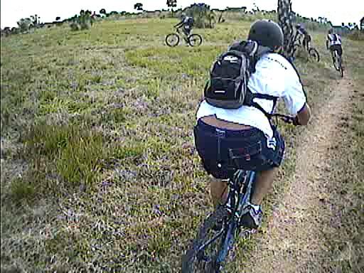 A group of mountain bikers riding along a grassy trail, with one cyclist in the foreground wearing a black helmet and a white shirt. The trail is surrounded by sparse vegetation and trees. Caloosahatchee Regional Park mountain bike trail.