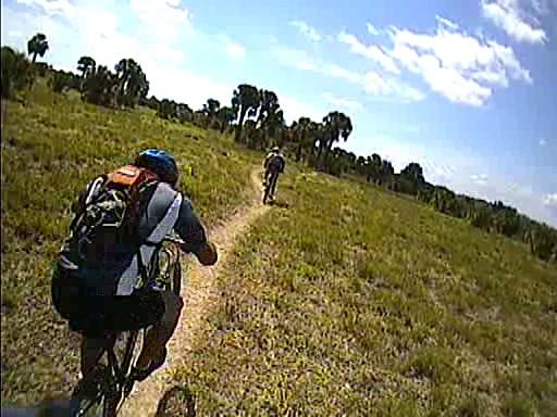 Two cyclists riding on a dirt path through a grassy field, surrounded by trees under a sunny sky. One cyclist is in the foreground, leaning forward, while the other cyclist is further ahead in the background. Caloosahatchee Regional Park mountain bike trail.
