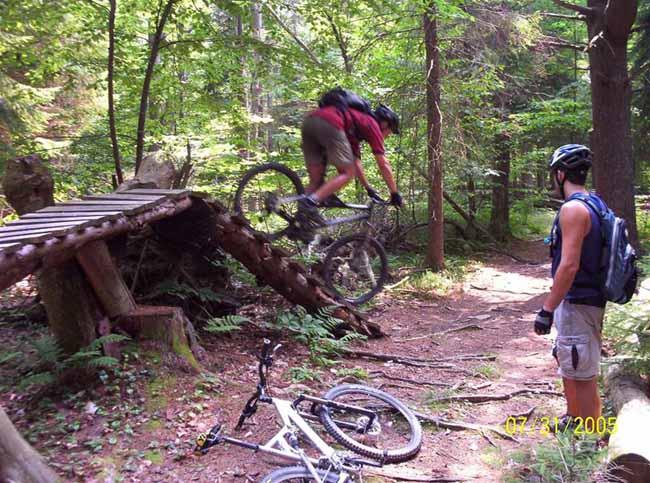A mountain biker jumps off a wooden ramp in a forest trail, while another cyclist watches nearby. The scene is surrounded by lush greenery, and two bikes are positioned on the ground. The action captures the excitement of mountain biking in a natural setting. Shindagin Hollow mountain bike trail.