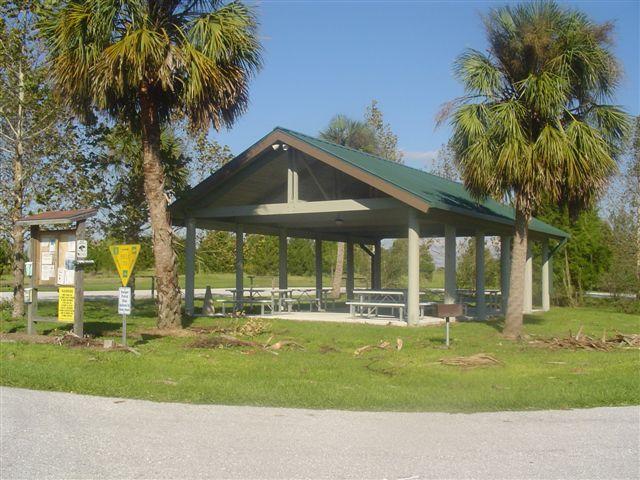 A covered picnic shelter with a green metal roof, surrounded by palm trees and grassy areas. The shelter features several picnic tables underneath. Nearby signage indicates park regulations and information, and a dirt road leads up to the shelter. Caloosahatchee Regional Park mountain bike trail.