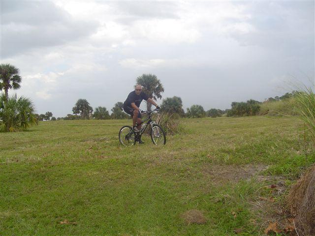 A person riding a mountain bike on a grassy field with palm trees in the background under a cloudy sky. Caloosahatchee Regional Park mountain bike trail.
