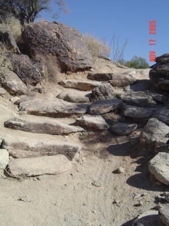A rocky path leading uphill, featuring uneven stone steps and surrounding dry terrain under a clear blue sky. South Mountain Park / National Trail mountain bike trail.